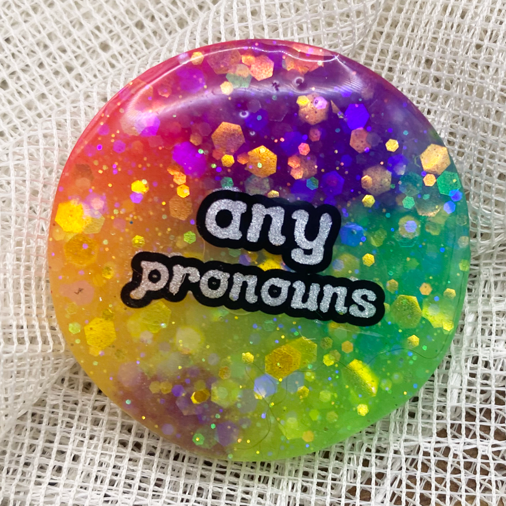 A glow in in the dark rainbow pronoun badge with any pronouns label