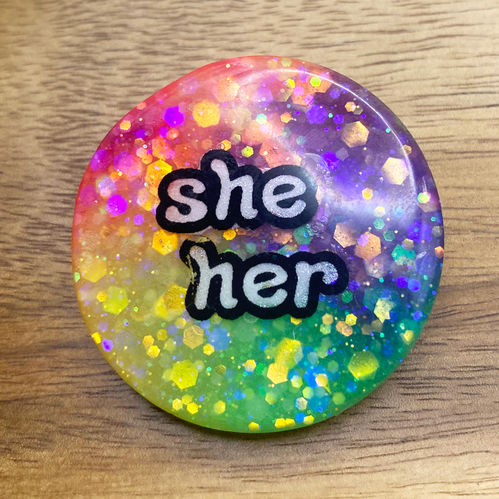 A glow in in the dark rainbow pronoun badge with she/her label - the badge is slightly discounted because of an imperfection on the label