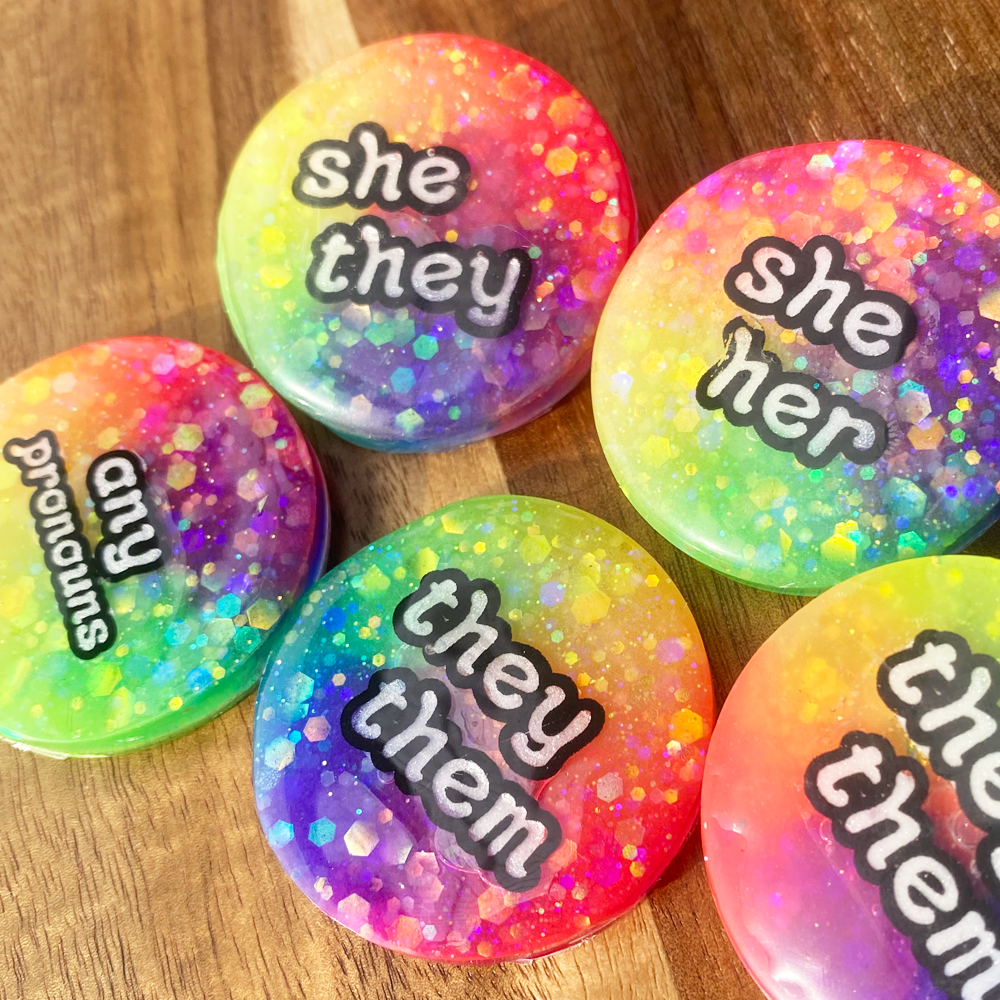 A collection of glow in in the dark rainbow pronoun badges - includes she/her, she/they, they/them and any pronouns options
