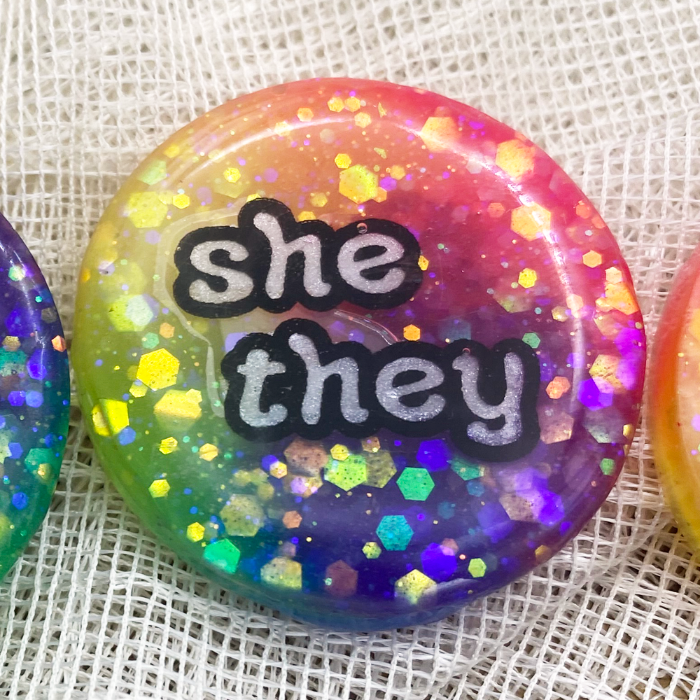 A glow in in the dark rainbow pronoun badge with she/they label