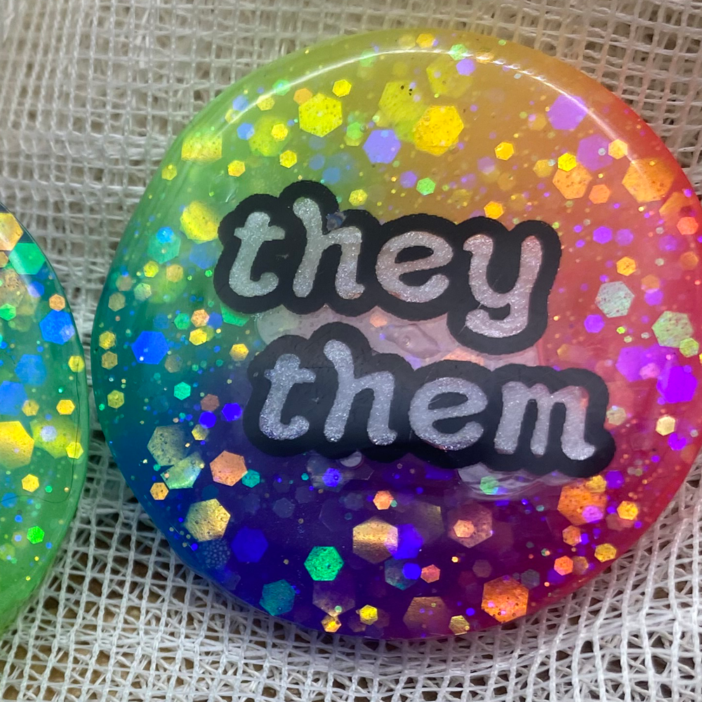 A glow in in the dark rainbow pronoun badge with they/them label