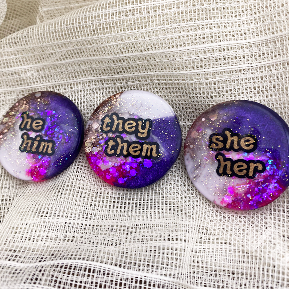Purple, pink, white and gold statement pronoun badges with she/her, they/them and he/him labels