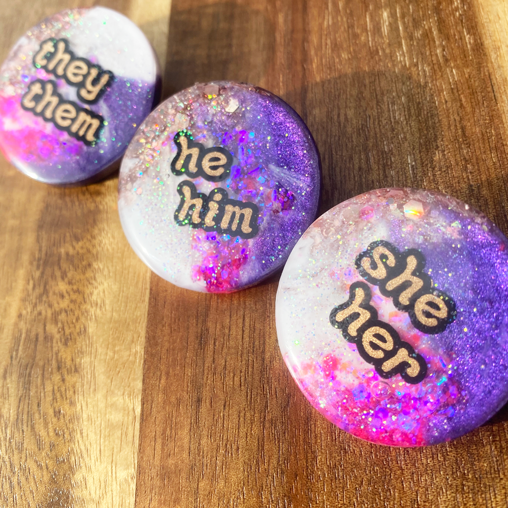 Purple, pink, white and gold statement pronoun badges with she/her, they/them and he/him labels in outdoor light on a wooden surface