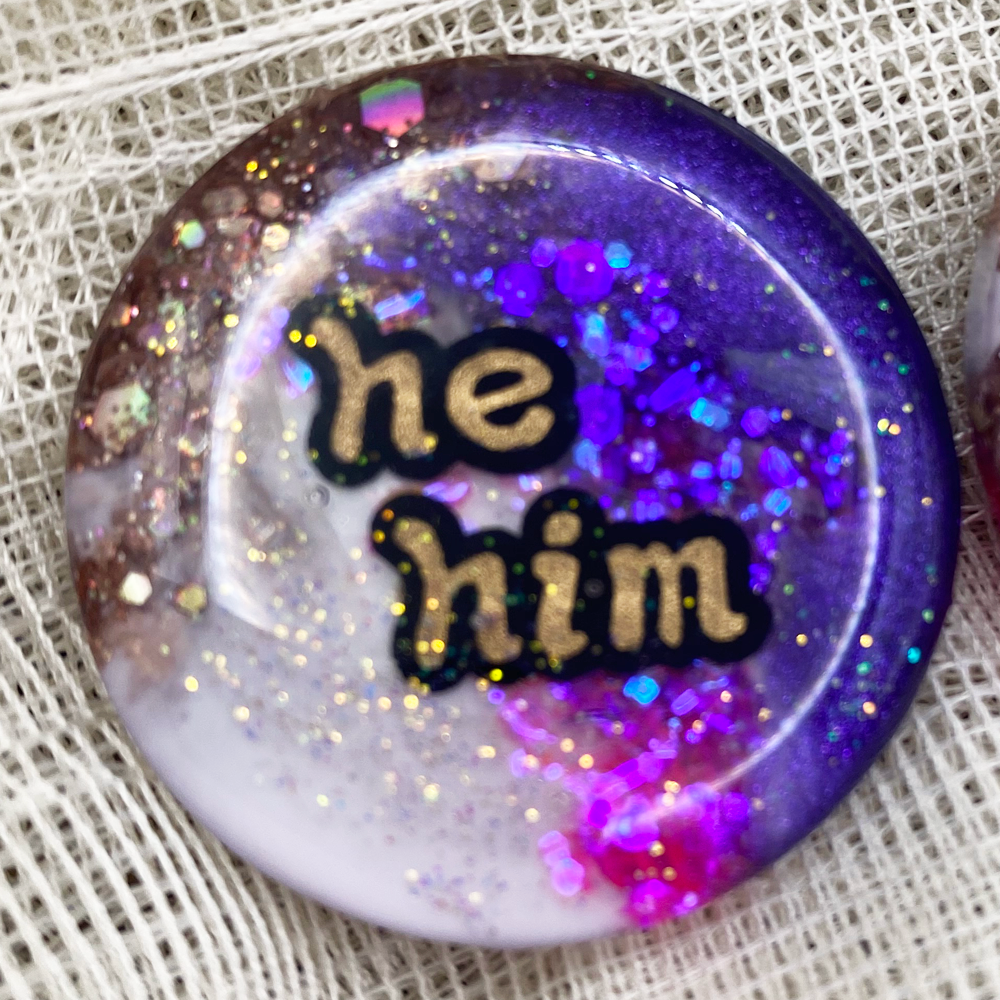Purple, pink, white and gold statement pronoun badge with  he/him label
