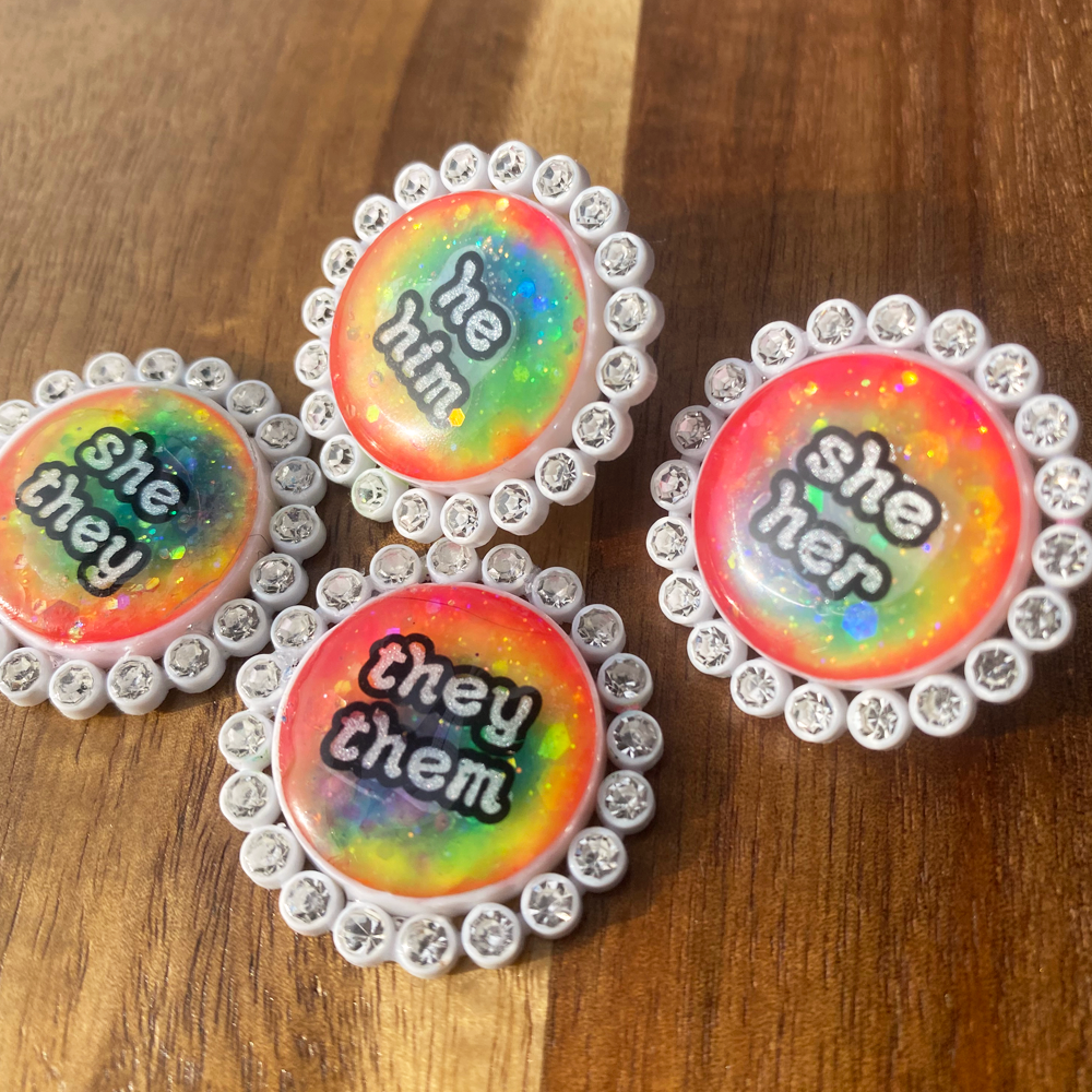 Four white rhinestone glowing rainbow pronoun pins with she/her, they/them and he/him labels on a wooden background