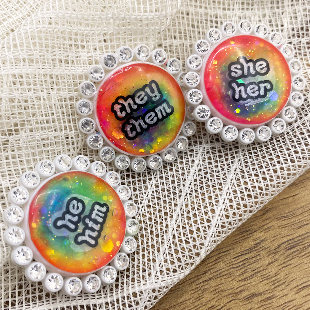 Three white rhinestone glowing rainbow pronoun pins with she/her, they/them and he/him labels
