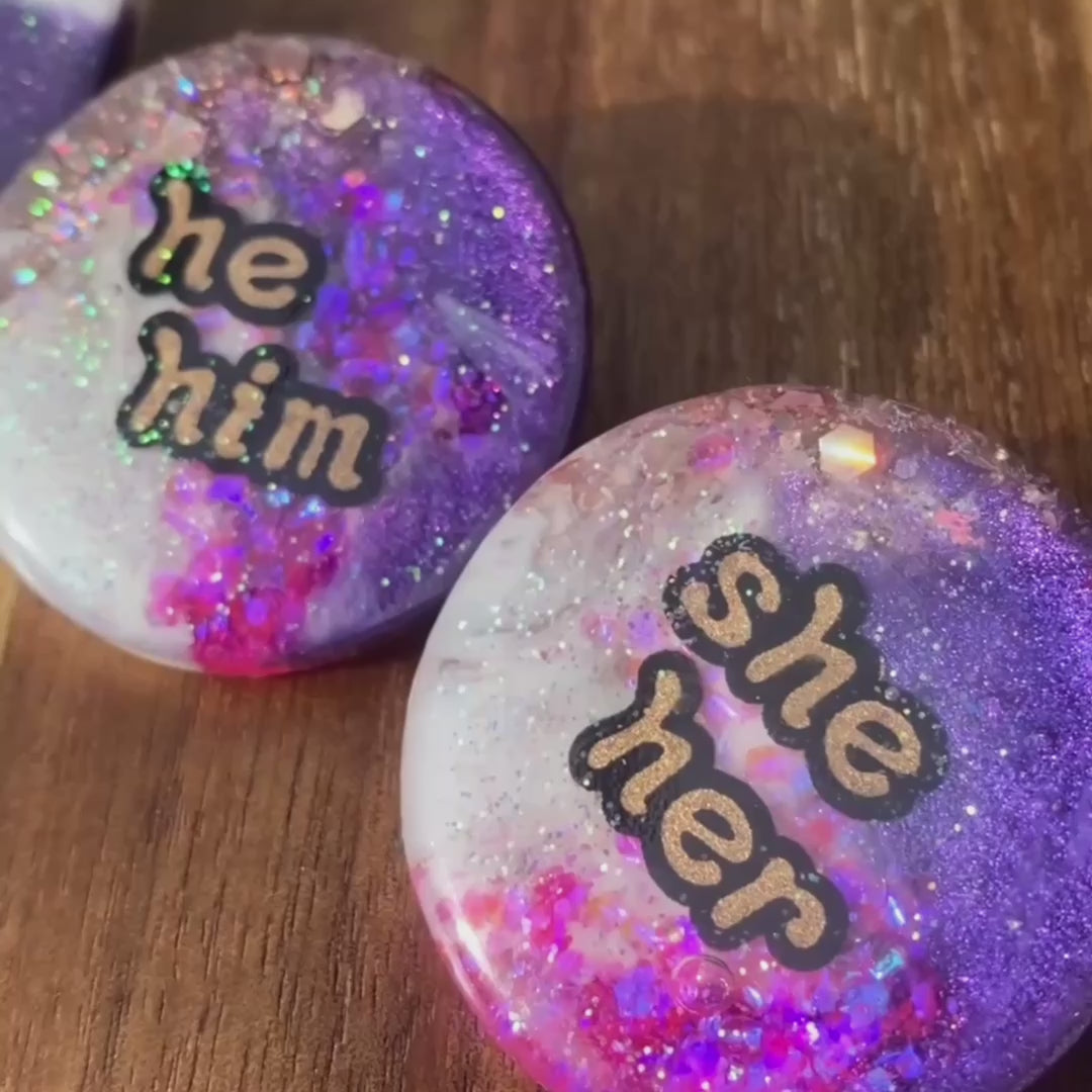 The video pans across these purple, pink, white and gold statement pronoun badges with she/her, they/them and he/him labels. the pronoun badges are handmade in Melbourne.