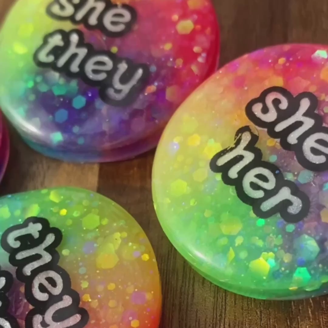 Various pans across rainbow pronoun badges on a wooden surface, mixed with footage of what these glow in the dark badges look like with the lights off