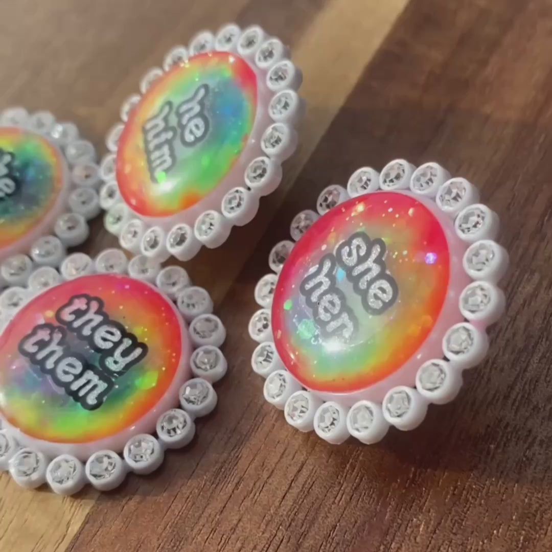The video pans across a collection of white rhinestone glowing rainbow pronoun pins with she/her, they/them and he/him labels. It shows the pins in daylight as well as the glow in the dark effect.