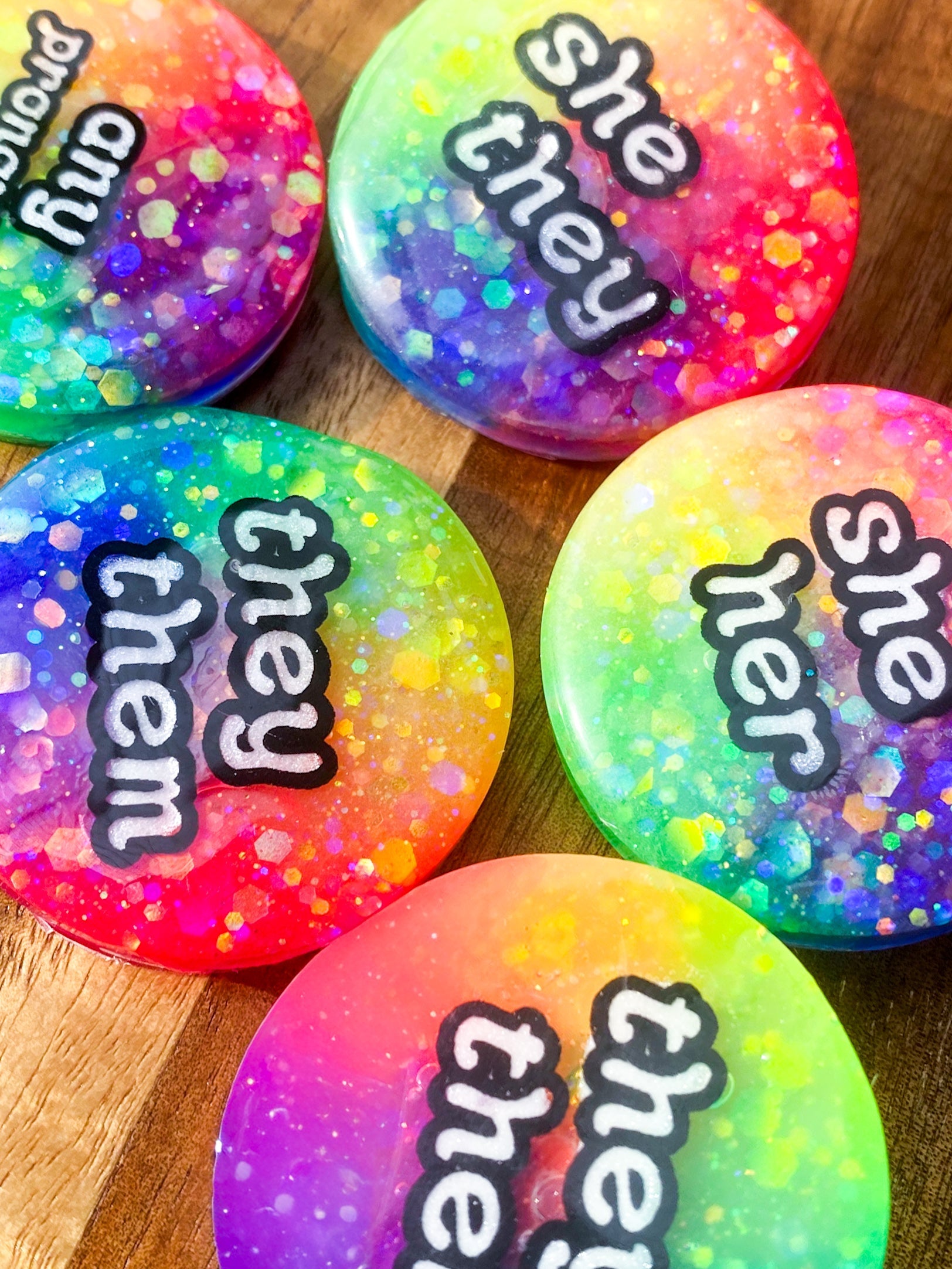 A collection of colourful, glittery resin pronoun badges, handmade in Melbourne