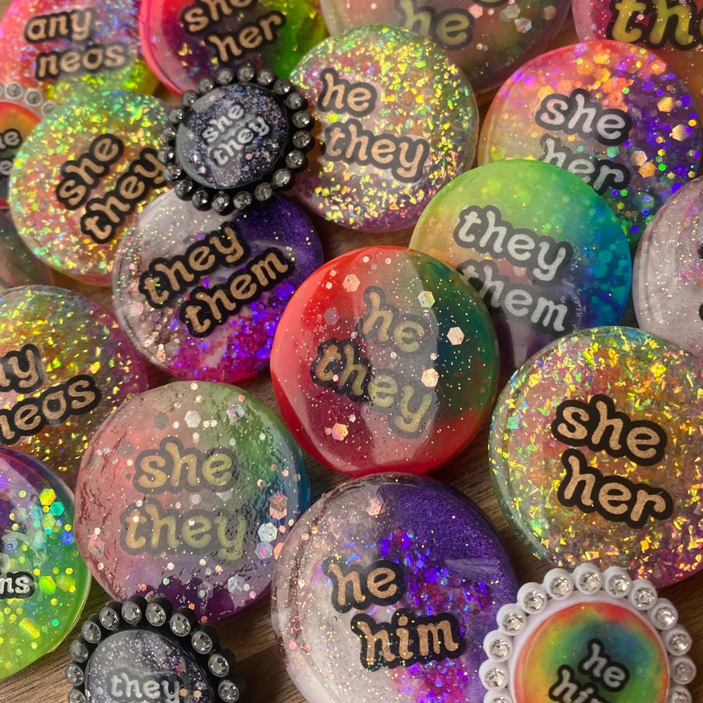 A collection of colourful, glittery resin pronoun badges, handmade in Melbourne