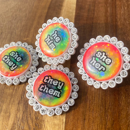 Four white rhinestone glowing rainbow pronoun pins with she/her, they/them and he/him labels on a wooden background