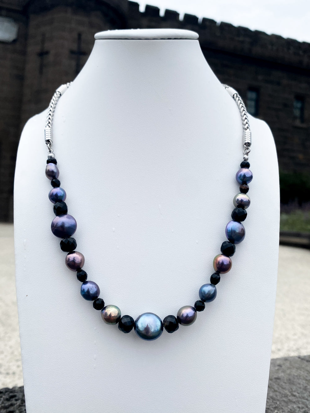 Beaded vibrant pearl and black agate necklace on white bust with shadowed bricks in background